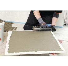 Eine Person trägt mit einer Zahnspachtel Kleber auf eine Fliese auf.