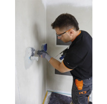 Ein Mann mit Schutzbrille arbeitet mit einer Spachtel an einer Wand im Badezimmer.