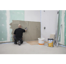 Ein Fliesenleger trägt Fliesenkleber auf eine Wand auf. Im Raum befinden sich ein Eimer mit Fliesenkleber und eine Wasserwaage.