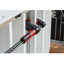 BESSEY Logo. Eine Schraubzwinge fixiert zwei Werkstücke.