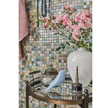 Dekoratives Arrangement mit Blumen, Vogel, Teekanne und Flakon auf einem Beistelltisch vor einer Mosaikwand