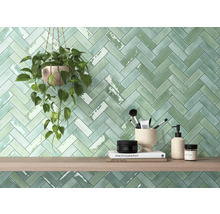 Wandgestaltung mit grünen, rechteckigen Fliesen im Fischgrätenmuster, Pflanze im Topf und Dekorationsartikeln auf einem Holzregal.