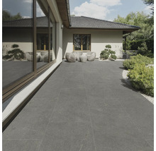 Terrasse mit grauen Fliesen, Gartenmöbeln und Bepflanzung vor einem Wohnhaus.