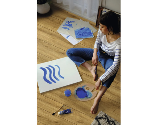 Eine Frau sitzt auf einem Laminatboden und malt mit blauer Farbe.