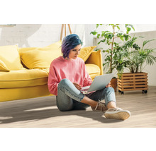 Frau sitzt auf dem Boden und benutzt einen Laptop, im Hintergrund Sofa und Pflanzen.