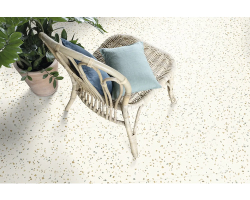 Heller Boden mit Terrazzo-Optik, ein Rattan-Stuhl mit Kissen und eine Zimmerpflanze