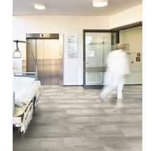 Grauer Bodenbelag in Steinoptik in einem hellen Krankenhausflur mit Patientenbett, Aufzug und Glastür.