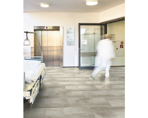 Grauer Bodenbelag in Steinoptik in einem hellen Krankenhausflur mit Patientenbett, Aufzug und Glastür.