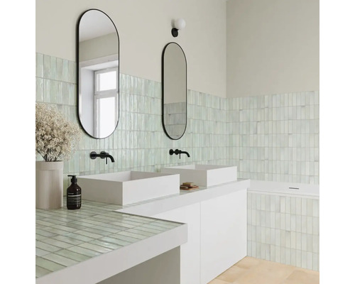 Helles Badezimmer mit zwei weißen Aufsatzwaschbecken, ovalen Spiegeln, schwarzen Armaturen und hellgrünen vertikalen Wandfliesen.