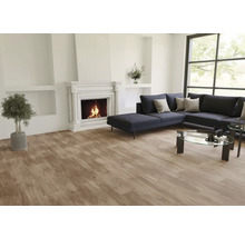 Modernes Wohnzimmer mit Vinylboden in Holzoptik, weißem Kamin mit brennendem Feuer, dunkelgrauem Sofa und Glastisch.