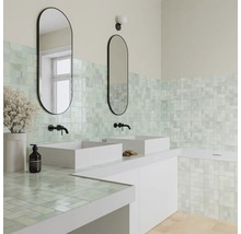Helles Badezimmer mit Mosaikfliesen an Wand und Arbeitsplatte, zwei weißen Aufsatzwaschbecken, schwarzen Armaturen und ovalen Spiegeln.