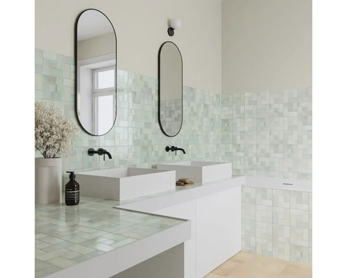 Helles Badezimmer mit Mosaikfliesen an Wand und Arbeitsplatte, zwei weißen Aufsatzwaschbecken, schwarzen Armaturen und ovalen Spiegeln.
