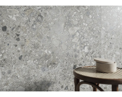 Wandfliesen in Terrazzo-Optik mit grauen und weißen Steineinschlüssen. Im Vordergrund ein runder Hocker mit Geflechtsitz und eine Keramikdose.