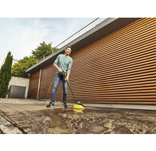 Ein Mann reinigt eine Steinterrasse mit einem Hochdruckreiniger und Flächenreiniger vor einer Holzfassade.