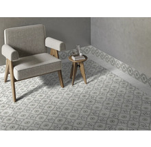 Hell gestalteter Raum mit gemusterten Bodenfliesen und passender Wandbordüre, ein grauer Sessel und ein runder Beistelltisch aus Holz.