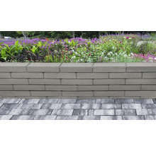 Gartenmauer aus glatten, grauen Betonsteinen vor einem bepflanzten Beet mit Blumen und Gräsern, angrenzend an eine gepflasterte Fläche.
