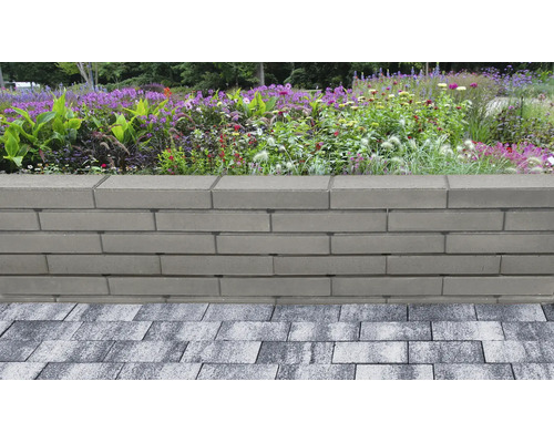 Gartenmauer aus glatten, grauen Betonsteinen vor einem bepflanzten Beet mit Blumen und Gräsern, angrenzend an eine gepflasterte Fläche.