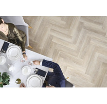 Draufsicht auf ein Esszimmer mit hellem Parkettboden in Fischgrätoptik, an einem gedeckten Tisch sitzen zwei Personen in eleganter Kleidung.
