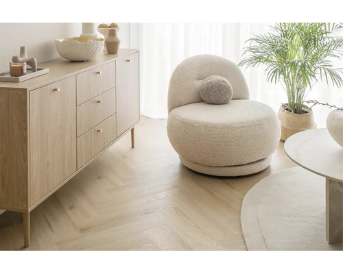 Helles Wohnzimmer mit einem beigen runden Sessel, einer Sideboard Kommode aus Holz und Fischgrätparkett.