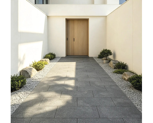 Moderner Eingangsbereich mit einem Gehweg aus großen grauen Terrassenplatten, gesäumt von weißem Kies, kleinen Sträuchern und Steinen.