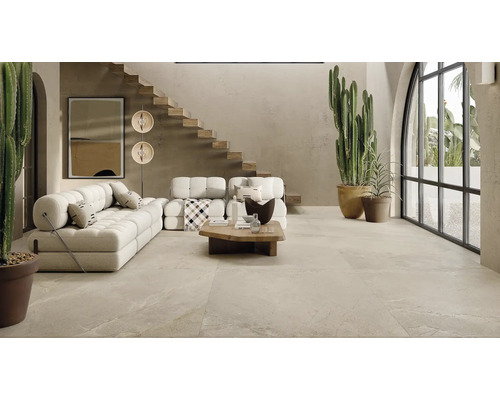 Geräumiges Wohnzimmer mit Steinfliesenboden, modernen Polstermöbeln, offener Holztreppe und großen Kakteen in einem hellen, naturnahen Interieur.