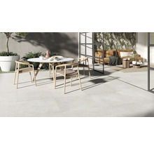 Moderne Terrasse mit hellen Bodenfliesen, runder Tischgruppe und Übergang in einen lichtdurchfluteten Wohnbereich mit raumhoher Verglasung.