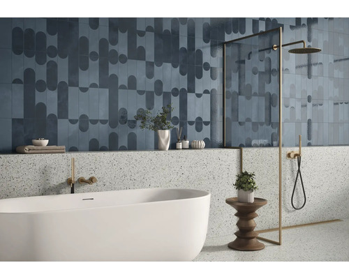 Modernes Badezimmer mit gemusterten Wandfliesen, Terrazzooptik im unteren Bereich, freistehender Badewanne und goldfarbener Duscharmatur.
