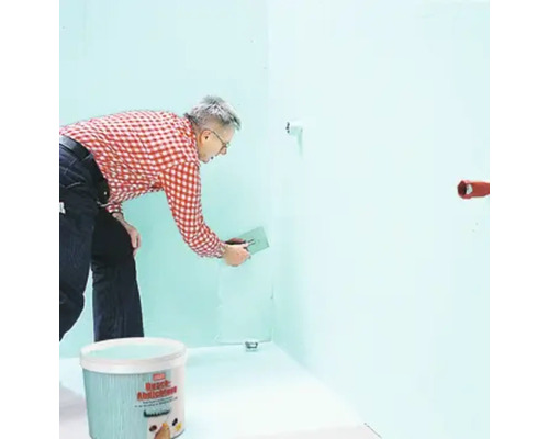 Ein Mann trägt Spachtelmasse auf eine Wand auf, im Vordergrund ein Eimer mit Spachtelmasse