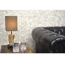 Szene mit einer Tischlampe auf einem Holztisch, zwei Gläsern, einem Flaschenöffner und einem Ledersofa vor einer Wand mit Kieselsteinen.