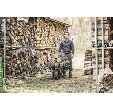 Mann transportiert Holzscheite mit einer Schubkarre vor einem Holzstapel.