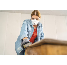 Frau schleift mit Schleifpapier ein Möbelstück, dabei trägt sie eine Schutzmaske und eine Schutzbrille