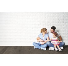 Familie sitzt auf einem dunklen Holzboden vor einer hellen Backsteinwand.