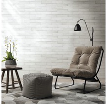 Wohnzimmer-Szene mit Sessel, Fußhocker, Beistelltisch, Stehlampe und Dekoration auf einem gemusterten Teppich vor einer Steinwand.