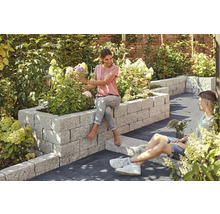 Garten mit Hochbeet aus Stein, bepflanzt mit Blumen, auf dem eine Frau sitzt, während ein Mann daneben auf dem Boden sitzt