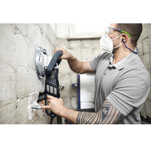 Mann arbeitet mit einer Mauernutfräse von Festool an einer Wand im Badezimmer. Er trägt eine Schutzmaske und eine Schutzbrille.