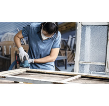 Frau schleift Fensterrahmen mit einem elektrischen Schleifer in einer Werkstatt.