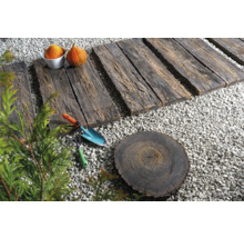 Gartenszene mit Holzwegplatten, Kies, Baumstumpfscheibe, Schaufel, Harke und Kürbissen.