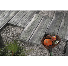 Gartenszene mit Gehwegplatten in Holzoptik, Zierkies und einem Korb mit Gartenutensilien.