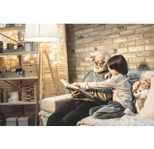 Ein Großvater liest mit seinem Enkel ein Buch in einem gemütlichen Raum mit Regalen, einer Stehlampe und einer Ziegelwand.