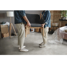 Zwei Personen tragen einen grauen Sessel mit Holzbeinen in einem Raum.