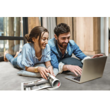 Ein Paar liegt auf einem Teppich und schaut auf einen Laptop mit einer Zeitschrift daneben.