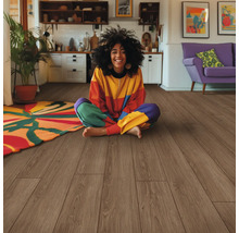 Eine Frau sitzt auf einem Holzboden in einem hellen Wohnraum mit einer Küche und einem Sofa im Hintergrund.