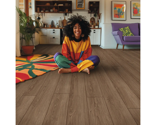 Eine Frau sitzt auf einem Holzboden in einem hellen Wohnraum mit einer Küche und einem Sofa im Hintergrund.