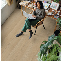 Frau sitzt mit Laptop auf einem Stuhl in einem Zimmer mit hellem Holzboden.