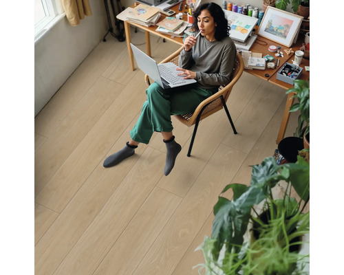 Frau sitzt mit Laptop auf einem Stuhl in einem Zimmer mit hellem Holzboden.