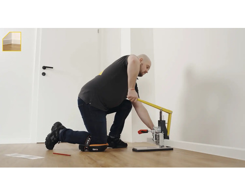 Ein Mann kniet auf einem Holzboden und verwendet ein Hebelwerkzeug zur Montage einer weißen Sockelleiste.