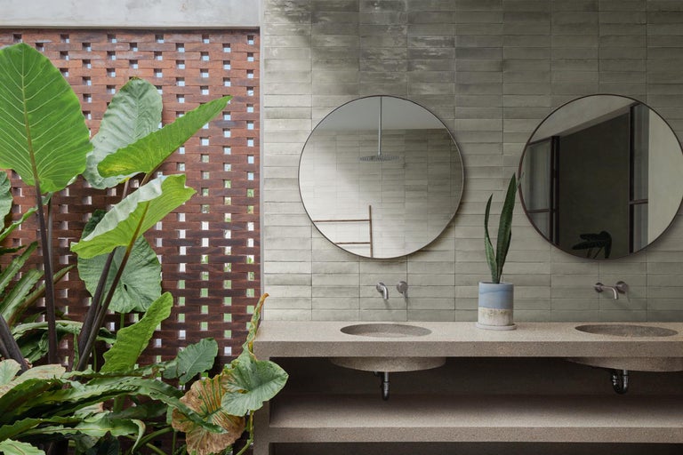 Modernes Badezimmer mit Doppelwaschtisch aus Stein, zwei runden Spiegeln, grauen Wandfliesen und großen tropischen Pflanzen.