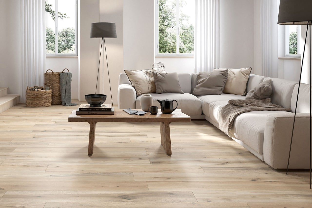 Helles, modernes Wohnzimmer mit hellem Bodenbelag in Holzoptik, einem Ecksofa, einem massiven Holztisch und großen Fenstern.