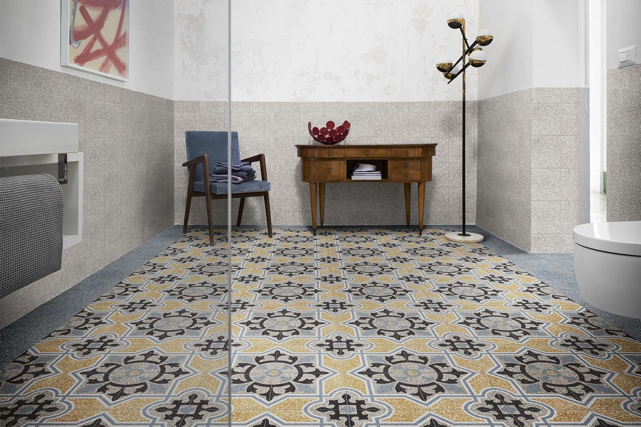 Badezimmer mit gemusterten Fliesen im Retro Stil und Terrazzo Wandfliesen, ergänzt durch einen antiken Holztisch und einen Stuhl.