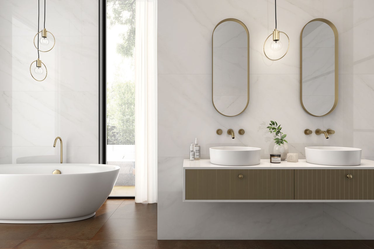 Helles, modernes Badezimmer mit Wänden in Marmoroptik, freistehender Badewanne, Doppelwaschtisch, goldenen Armaturen und ovalen Spiegeln.